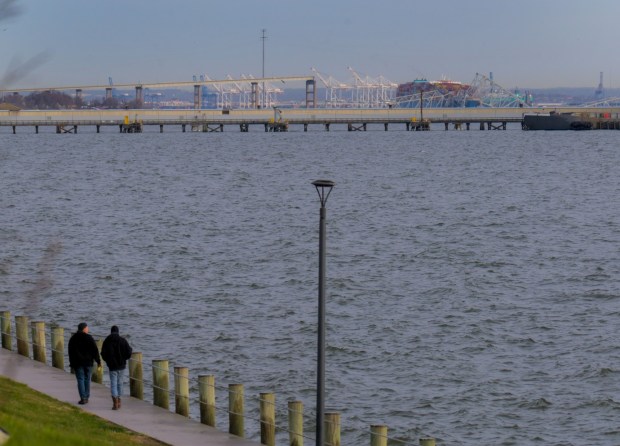 People walk along a pier as the western span of the Francis Scott Key Bridge rises over the Patapsco River where a container ship, back right collided with the structure overnight. (Karl Merton Ferron/Staff)